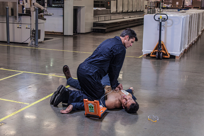 Man performing CPR on a warehouse co-worker with an AED at his side, pads attached.