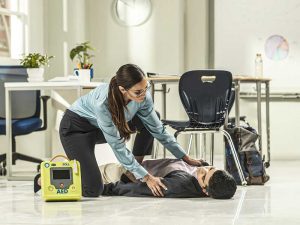 Woman in classroom checking an unresponsive man on the floor with an AED at her side.