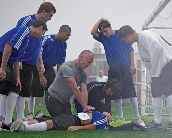 Group of high school soccer players huddle around coach performing CPR on a downed player suffering cardiac arrest using a ZOLL AED Plus.
